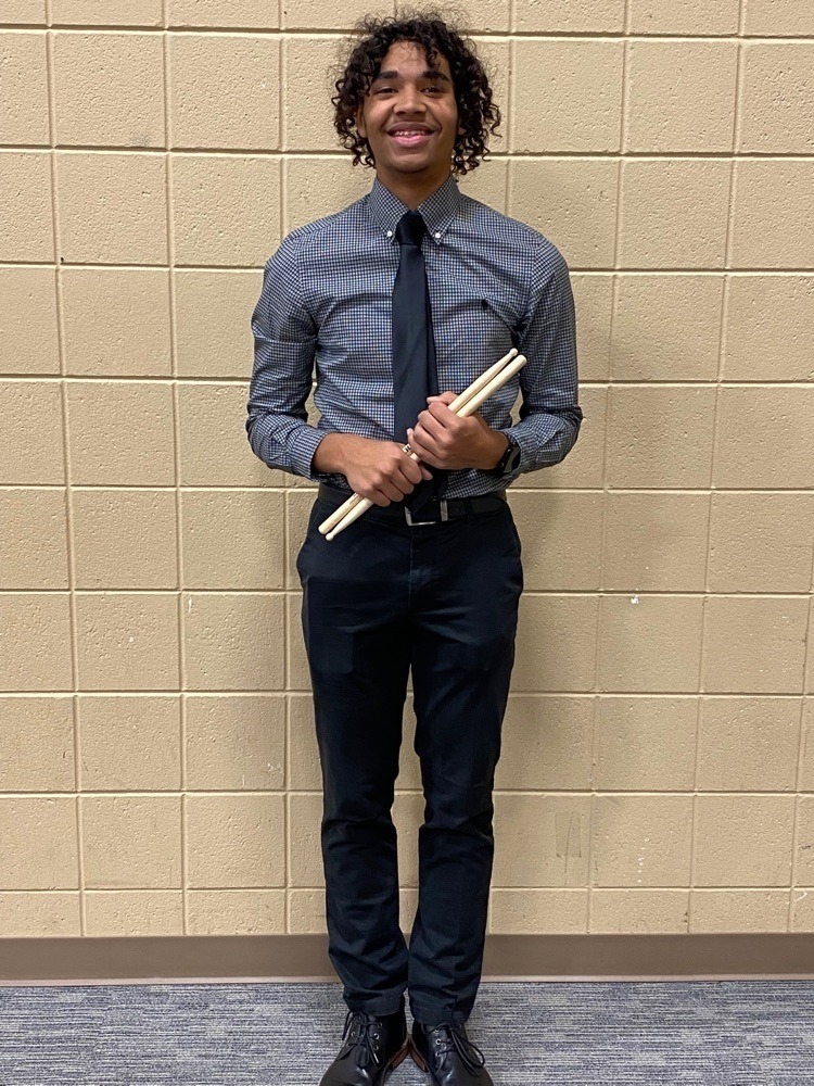 ​young man in dress shirt, dress pants, and a tie, holding drumsticks. He's standing in front of a beige background with blue carpeting​​