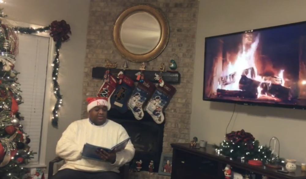 man in a santa hat in front of a fireplace with four stockings holding a book.  a tv displaying a fireplace is to the right of the image. decorated christmas tree is to the far left.