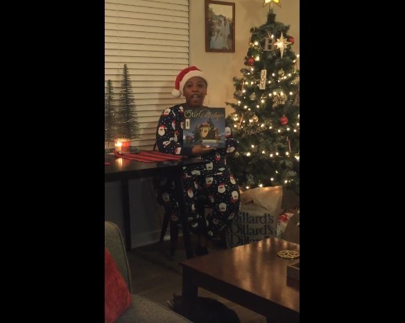 woman in santa pajamas and a santa hat in front of beige blinds holding a book. she's sitting to the left of a black table. On her left is a decorated christmas tree. There's a coffee table in front of her and a gray sofa in the foreground.